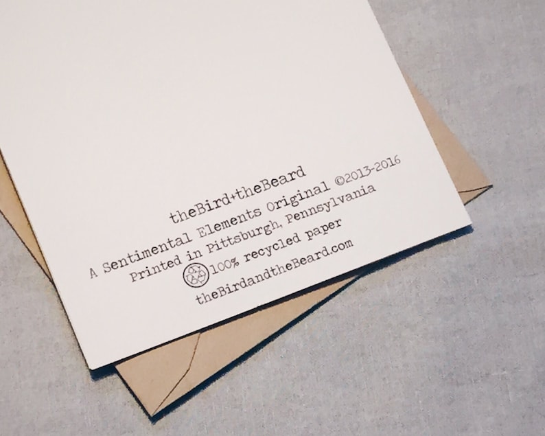 The back of a white greeting card reads theBird+theBeard, A Sentimental Elements Original. Printed in Pittsburgh, Pennsylvania., 100% recycled paper. A Kraft envelope lays behind the card. The background is gray.