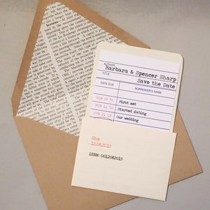 A library borrower’s card customized as a wedding save the date announcement, is placed inside library sleeve and sits atop a Kraft paper bag envelope that is lined with an encyclopedia page. The background is gray.