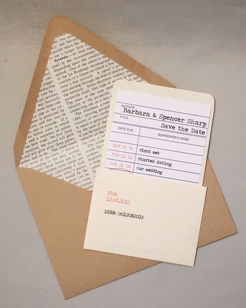 A library borrower’s card customized as a wedding save the date announcement, is placed inside library sleeve and sits atop a Kraft paper bag envelope that is lined with an encyclopedia page. The background is gray.