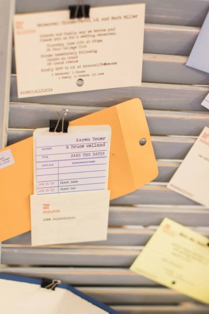 A library borrower’s card and sleeve, used as a wedding save the date announcement is clipped to a light blue shutter with a Kraft catalog envelope. Other literary themed invitations appear blurred out in the background.