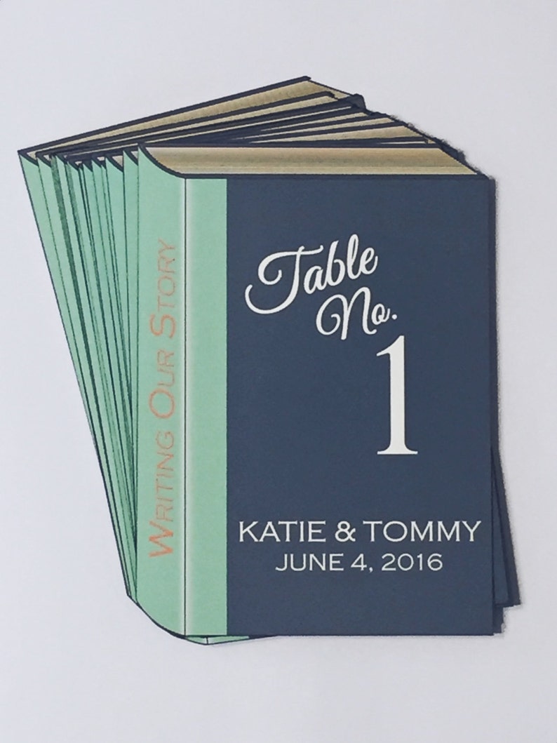 Puede incluir: Una pila de libros de color azul marino, turquesa y marr&oacute;n con una cubierta azul que dice "Table No. 1" y "Katie & Tommy June 4, 2016". El lomo de los libros dice "Writing Our Story".