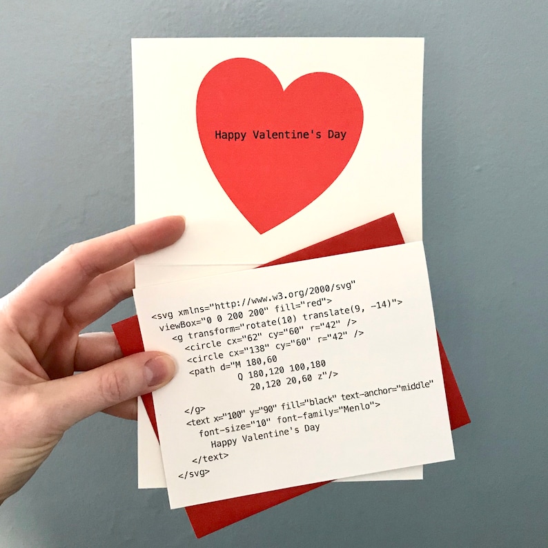 以下が含まれることがあります： 白いカードに赤いハートと「Happy Valentine's Day」の文字が書かれています。カードは半分に折り畳まれており、内側にはハートのSVG画像のコードスニペットが表示されています。