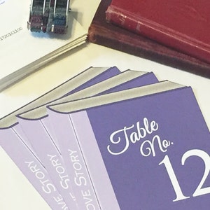 Puede incluir: Tarjeta de n&uacute;mero de mesa de color morado con el texto "Table No. 12" en letra blanca. La tarjeta est&aacute; dise&ntilde;ada para parecer una pila de libros con el texto "Love Story" en los lomos.