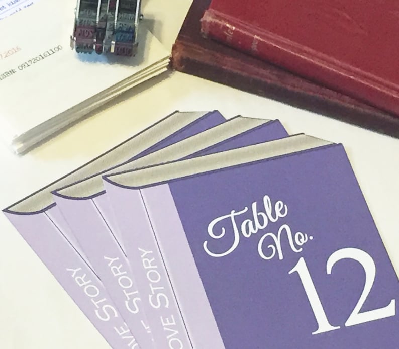 Puede incluir: Tarjeta de n&uacute;mero de mesa de color morado con el texto "Table No. 12" en letra blanca. La tarjeta est&aacute; dise&ntilde;ada para parecer una pila de libros con el texto "Love Story" en los lomos.