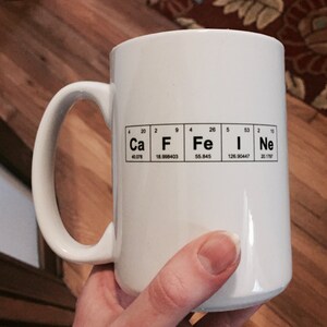 A hand holds a white mug with Caffeine spelled out using the Periodic Table of the elements. This image shows that the mug is an imperfect second as the design is printed slightly off center.