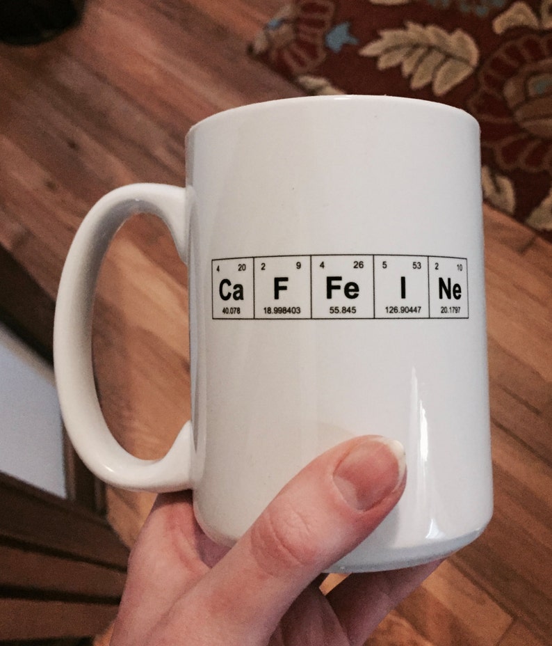 A hand holds a white mug with Caffeine spelled out using the Periodic Table of the elements. This image shows that the mug is an imperfect second as the design is printed slightly off center.