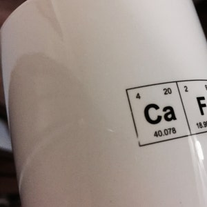 A detail image of a white mug with Caffeine spelled out using the Periodic Table of the elements. This image shows that the mug is an imperfect second as there are areas where the ink is light or missing in the design.