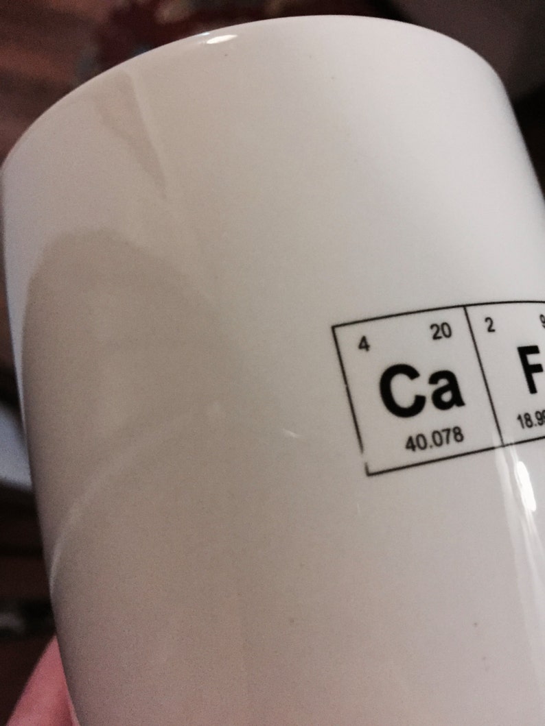 A detail image of a white mug with Caffeine spelled out using the Periodic Table of the elements. This image shows that the mug is an imperfect second as there are areas where the ink is light or missing in the design.