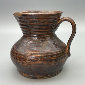 May include: A handcrafted brown ceramic pitcher with a curved handle. The pitcher has a wide base that tapers to a narrower neck and a flared rim. The surface is textured with horizontal lines, giving it a rustic appearance. The pitcher is suitable for serving beverages.