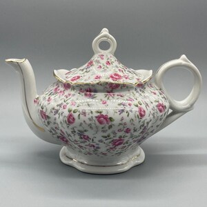 Puede incluir: Una tetera de porcelana blanca con un denso patrón floral de rosas rosadas, hojas verdes y pequeñas flores azules. La tetera tiene un borde festoneado, un asa curva, un pico vertedor y una tapa con un remate circular, todo acentuado con bordes dorados.