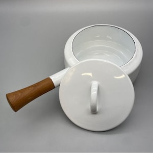 Puede incluir: Cacerola de esmalte blanco con mango de madera y tapa a juego. La cacerola tiene un borde negro y un asa blanca en la tapa. El mango de madera es de color marrón claro.