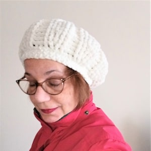May include: A white crocheted beret with a textured design. The beret is worn by a person with brown hair and glasses. The person is wearing a red jacket.