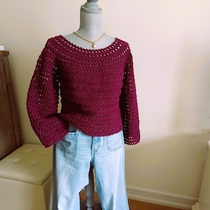 May include: A maroon crocheted sweater with bell sleeves. The sweater is on a mannequin and has a delicate gold chain necklace.