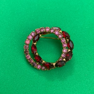 May include: A gold-toned brooch with a double circle design. The circles are set with red and pink rhinestones.