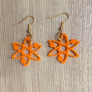 Może przedstawiać: Para pomarańczowych kolczyków w kształcie atomu ze złotymi haczykami. Kolczyki są w jasnym, jednolitym pomarańczowym kolorze i mają stylizowany wzór atomu z okrągłymi akcentami. Kolczyki są prezentowane na jasnym tle z usłojeniem drewna.