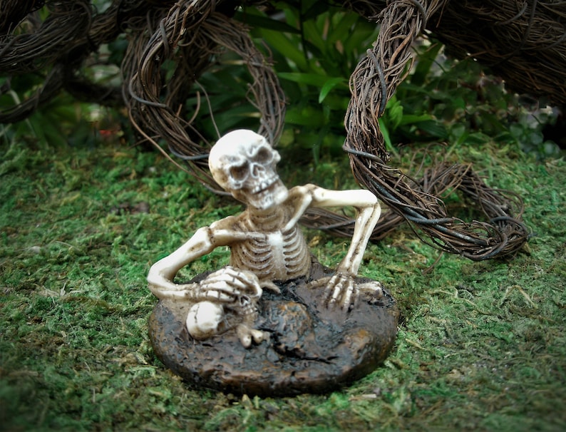 Peut inclure: Une figurine de squelette blanc est partiellement enterr&eacute;e dans le sol, avec ses bras et sa t&ecirc;te qui d&eacute;passent. Le squelette est pos&eacute; comme s'il rampait hors du sol. La figurine est entour&eacute;e de mousse verte et de vignes brunes.