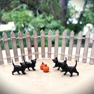May include: Four small black cat figurines standing in a semi-circle around two miniature orange pumpkins, positioned in front of a curved, weathered white wooden picket fence. The cats are depicted in various walking poses with tails raised.