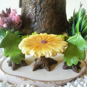 May include: A miniature fairy garden set featuring a yellow flower table with a purple center and two green leaf chairs. The table and chairs are set on a wooden base, surrounded by miniature plants and white pebbles.