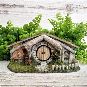May include: A miniature stone cottage with a round wooden door, a small window, and a picket fence. The cottage is surrounded by green foliage.