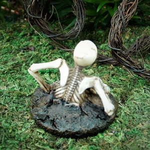 Peut inclure: Une figurine de squelette blanche est partiellement enterr&eacute;e dans un monticule de terre brune. Le squelette est pos&eacute; les bras lev&eacute;s et la t&ecirc;te renvers&eacute;e. La figurine est entour&eacute;e de mousse verte.