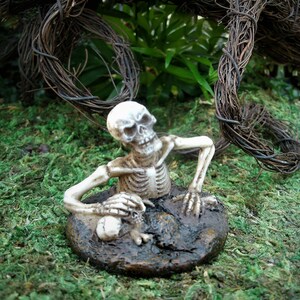 Peut inclure: Une figurine de squelette blanc est pos&eacute;e en position assise, les bras et les jambes pli&eacute;s. Le squelette est assis sur une base brune textur&eacute;e qui ressemble &agrave; de la terre. La figurine est entour&eacute;e de mousse verte.