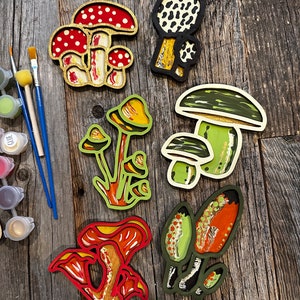 Op de afbeelding: Zes houten paddenstoelversieringen, geschilderd in verschillende kleuren en patronen. De paddenstoelen zijn allemaal van verschillende vormen en maten, en sommige hebben witte vlekken. De versieringen zijn gerangschikt op een houten oppervlak.