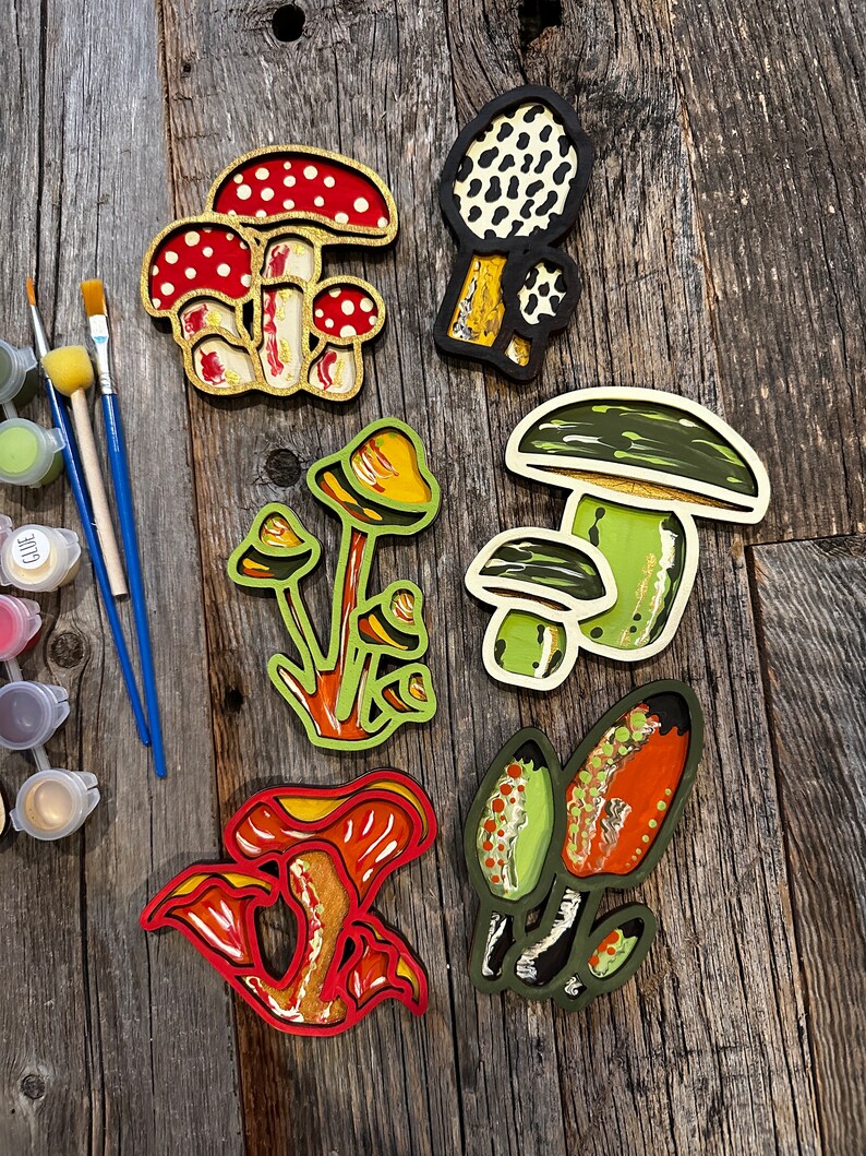 Op de afbeelding: Zes houten paddenstoelversieringen, geschilderd in verschillende kleuren en patronen. De paddenstoelen zijn allemaal van verschillende vormen en maten, en sommige hebben witte vlekken. De versieringen zijn gerangschikt op een houten oppervlak.