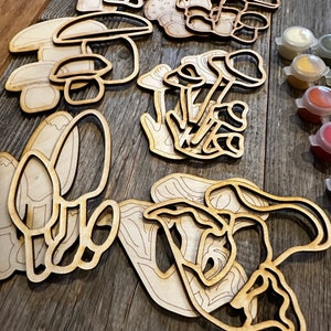 Op de afbeelding: Een set houten paddenstoelvormen om te beschilderen. De paddenstoelen zijn in verschillende maten en vormen, en sommige hebben details zoals stelen en hoeden. De set wordt geleverd met een verscheidenheid aan verfkleuren.