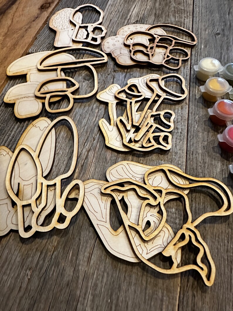 Op de afbeelding: Een set houten paddenstoelvormen om te beschilderen. De paddenstoelen zijn in verschillende maten en vormen, en sommige hebben details zoals stelen en hoeden. De set wordt geleverd met een verscheidenheid aan verfkleuren.