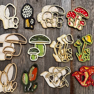 Op de afbeelding: Een set van 12 houten paddenstoelversieringen in verschillende vormen, maten en kleuren. De paddenstoelen zijn geschilderd in tinten bruin, wit, rood, groen en geel. Ze zijn perfect om een vleugje whimsy toe te voegen aan je interieur.