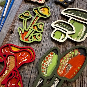 Op de afbeelding: Een set van zes houten paddenstoelversieringen, elk geschilderd in een andere kleur en patroon. De paddenstoelen zijn gelaagd en hebben een 3D-effect. De versieringen zijn perfect om elke inrichting een vleugje whimsy te geven.