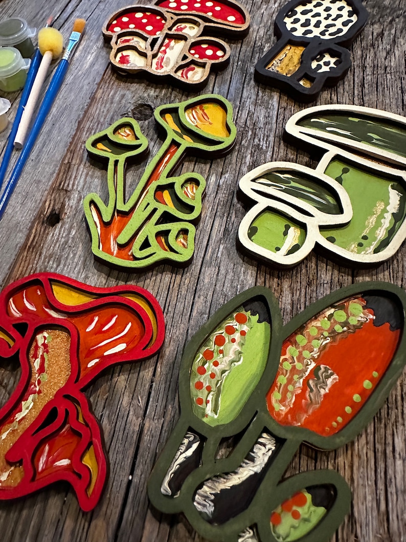 Op de afbeelding: Een set van zes houten paddenstoelversieringen, elk geschilderd in een andere kleur en patroon. De paddenstoelen zijn gelaagd en hebben een 3D-effect. De versieringen zijn perfect om elke inrichting een vleugje whimsy te geven.