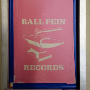 May include: A pink screen print with the words "BALL PEIN RECORDS" in white, along with a graphic of an anvil and hammer. The print is framed in a silver metal frame with a blue border.
