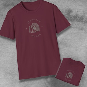 Op de afbeelding: Bordeauxkleurig T-shirt met de tekst "YEARN FOR THE URN" in een cirkelvormig ontwerp. Het ontwerp bevat een afbeelding van een treurwilg en een decoratieve urn. Een tweede T-shirt is zichtbaar in de afbeelding.