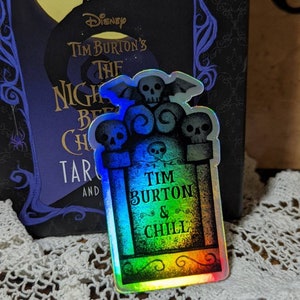 May include: A holographic sticker in the shape of a tombstone with the text "Tim Burton & Chill." The sticker features three skulls and a bat. The background includes a book with the title "Tim Burton's The Nightmare Before Christmas."