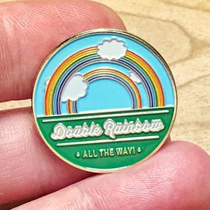 May include: A round enamel pin with a double rainbow design. The pin features a blue sky, white clouds, and a green banner with the words "Double Rainbow ALL THE WAY!" in white lettering. The pin has a gold-colored border.