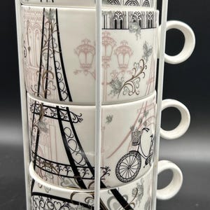 May include: A set of four white ceramic teacups with handles, stacked in a white metal rack. Each cup features a different Parisian-themed design in black, gray, and pink, including the Eiffel Tower, Notre Dame, and bicycles. The cups are approximately 3 inches in diameter.