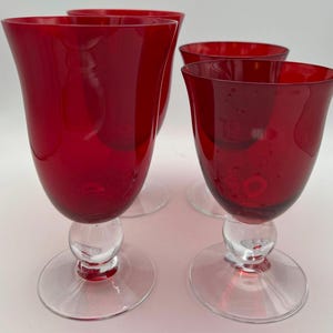 May include: A set of four drinking glasses featuring deep red bowls and clear glass stems with a spherical accent above a circular base. The glasses have a smooth, curved silhouette and a consistent design, showcasing a vibrant ruby color throughout.