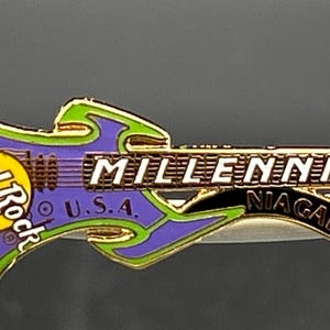 May include: A Hard Rock Cafe guitar-shaped pin. The pin is purple and green with gold trim. It reads "Hard Rock Cafe" and "Millennium Niagara Falls U.S.A."