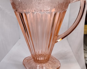 May include: A pink glass pitcher with a geometric design. The pitcher has a square base with a floral pattern, a flared body with vertical lines, and a decorative floral band near the top. It features a handle and a pouring spout.