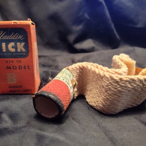May include: An orange box of Aladdin Wick next to a beige lamp wick. The box has blue text that reads "Aladdin WICK" and "USE IN MODEL 5". The lamp wick is beige and red with a black rim.
