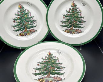 May include: Three white ceramic plates with a green border. Each plate features a detailed illustration of a Christmas tree decorated with ornaments and presents. The plates are arranged on a dark surface.