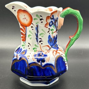 May include: A white ceramic pitcher with a hexagonal base, featuring hand-painted floral designs in orange, blue, and green. The handle is green with red accents. The pitcher has a scalloped rim and gold trim.