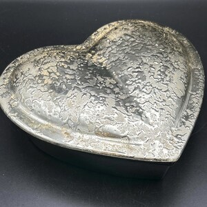 May include: A silver-colored heart-shaped trinket box with a textured surface. The box has a lid and a dark base. The surface of the lid has a mottled appearance, with areas of lighter and darker silver tones.