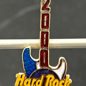 May include: A Hard Rock Cafe Ottawa souvenir pin in the shape of a guitar. The pin is primarily blue, red, and gold, with the year "2000" displayed vertically. The pin features the Hard Rock Cafe logo and the city name.