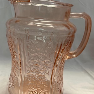 May include: A translucent light pink glass pitcher featuring a molded floral pattern of roses across its body. The pitcher has a wide spout, a curved handle on the right side, and a smooth, flared rim. The glass has a glossy finish throughout.