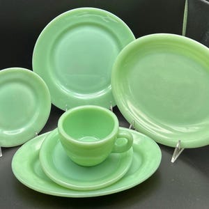Puede incluir: Una colección de vajilla verde jade, que incluye platos de varios tamaños, una taza y platillos. El juego incluye un plato pequeño, mediano y grande, y una fuente ovalada. El juego de taza y platillo está en el centro. Los platos se exhiben sobre soportes transparentes.