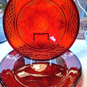May include: Two red glass plates with diamond and floral patterns. The plates are displayed on a clear stand, showcasing their intricate designs. The plates have a deep red color and are likely for serving or display.