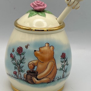 Może przedstawiać: Ceramiczny słoik na miód z ilustracją Kubusia Puchatka, różami i pszczołami. Pokrywka posiada uchwyt w kształcie wyrzeźbionej różowej róży oraz pasującą łyżeczkę z pszczołą na szczycie. Słoik ma złote wykończenia na krawędziach i podstawie.