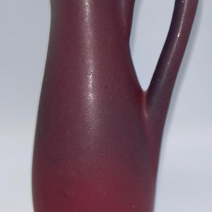 Puede incluir: Una jarra de cerámica de color burdeos con un asa curva y un pico acampanado. La jarra tiene un acabado liso y mate y una base redondeada. El asa es de un tono burdeos más oscuro. La jarra mide aproximadamente 18 cm de altura.